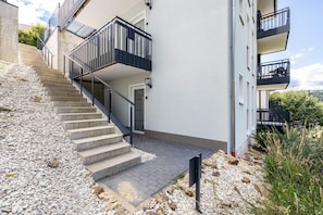 Un bâtiment moderne doté d’un escalier menant à un balcon, d’une zone pavée et offrant une vue sur la verdure.