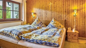 Ein Zimmer mit einer holzgetäfelten Wand, einem Bett mit floralem Bettzeug, einem Nachttisch mit Lampe und einem Fenster mit Blick ins Grüne.