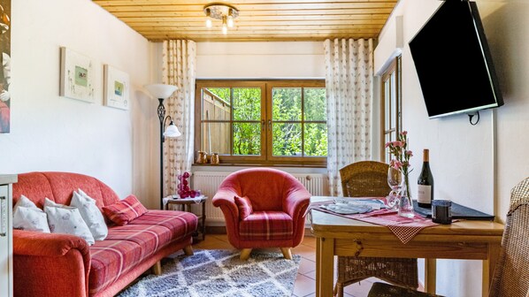 A cozy living room with a red sofa, a wooden table with a bottle and glass, and a large window with curtains.
