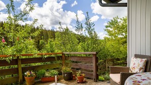A wooden deck with potted plants, a wicker chair, and a table with a floral cushion.