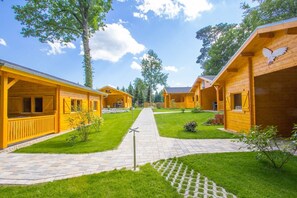Een rij houten hutten met een verhard pad ertussen, omringd door groen gras en bomen.