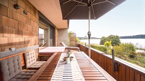 A balcony with wooden flooring, cushioned seating, a table with a striped tablecloth, and a view of a lake. - Apartment "Seen-Sucht" with Lake View, Shared Garden & Wi-Fi (Bad Endorf)