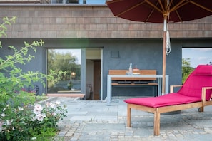 Un área de estar moderna al aire libre con una silla reclinable de madera y una mesa, rodeada por un jardín con plantas en flor.