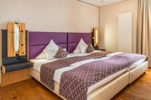 A modern hotel room with a large bed, a wooden side table, a wall-mounted light, and a plain wall.