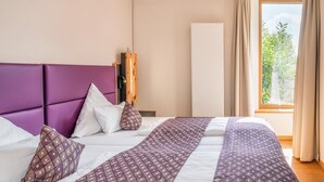 A hotel room with a bed, a wooden headboard, a window with curtains, and a view of greenery outside. - Apartment "Wellvital" with Private Terrace & Wi-Fi (Bad Endorf)
