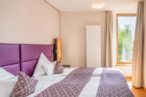 Habitación de hotel con cama, cabecero de madera, ventana con cortinas y vistas a zonas verdes.