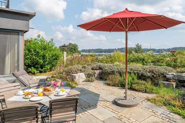 An outdoor patio with a table set for breakfast, a red umbrella, and a scenic view of a lake.