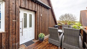 Een houten gevel met een witte deur, terras met rieten stoelen en een bloempot.