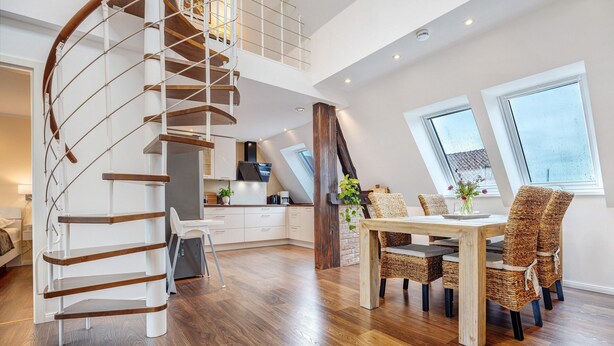 Een ruim appartement in loftstijl met een wenteltrap, een eethoek met rieten stoelen en een keuken met witte kasten.