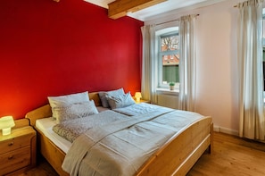 A bedroom with a wooden bed, a nightstand, a window with curtains, and a view of a building outside.