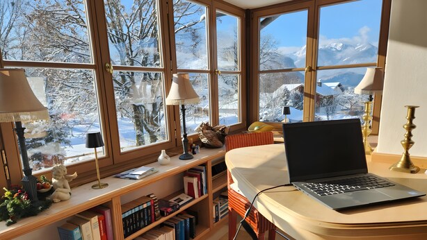 A cozy room with a laptop on a desk, a bookshelf filled with books, and large windows offering a snowy mountain view. - Spacious Group Holiday Home in Chiemgau – Ideal for Families & Friends (up to 10 guests) (Unterwössen)