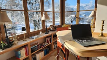 Une pièce chaleureuse avec un ordinateur portable posé sur un bureau, une bibliothèque remplie de livres et de grandes fenêtres donnant sur un paysage de montagne enneigé.