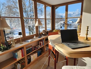 Une pièce chaleureuse avec un ordinateur portable posé sur un bureau, une bibliothèque remplie de livres et de grandes fenêtres donnant sur un paysage de montagne enneigé.