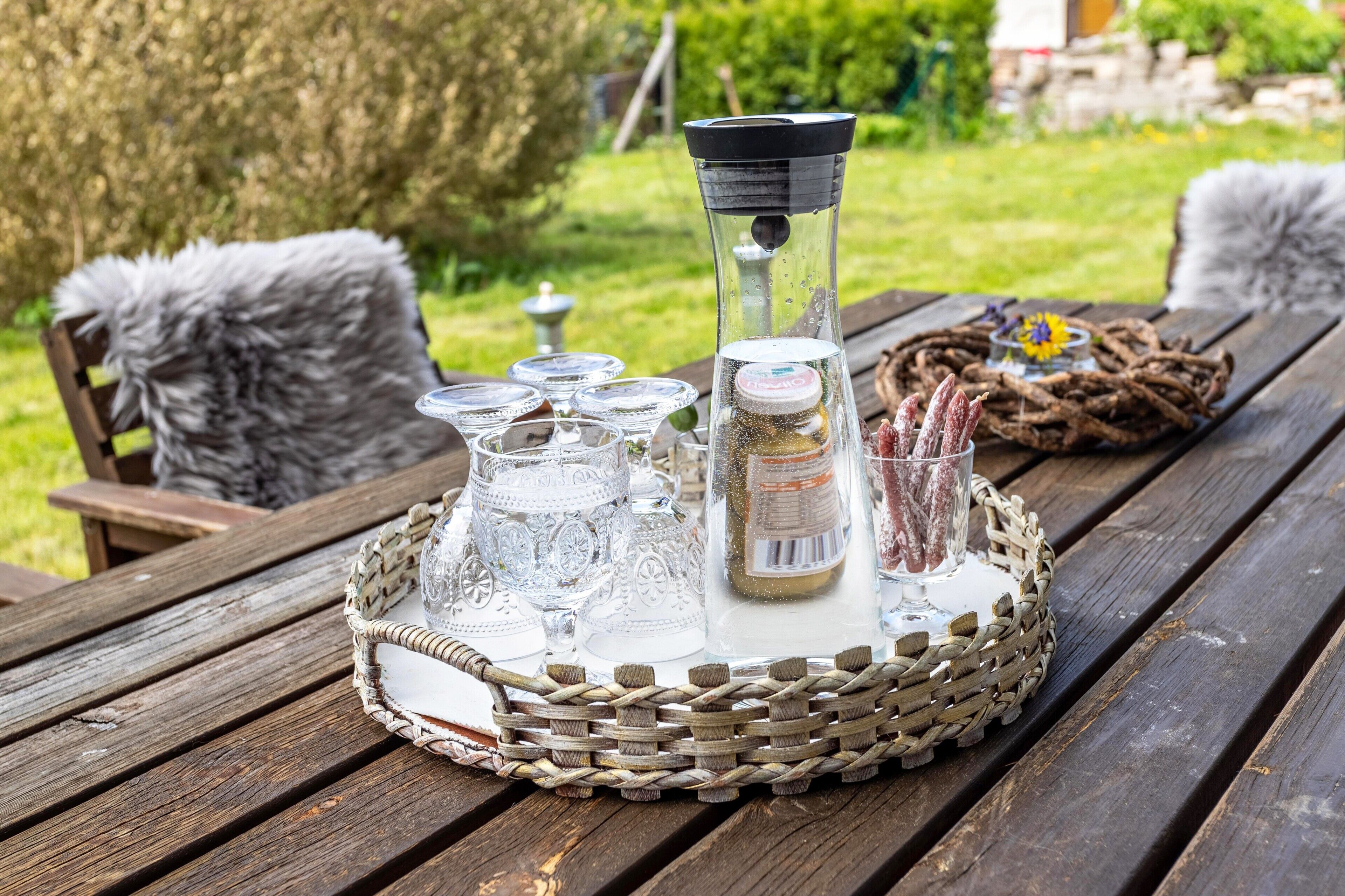 Una mesa de picnic de madera con una bandeja tejida que contiene cristalería, un dispensador de agua y una cesta con una vela y flores secas.
