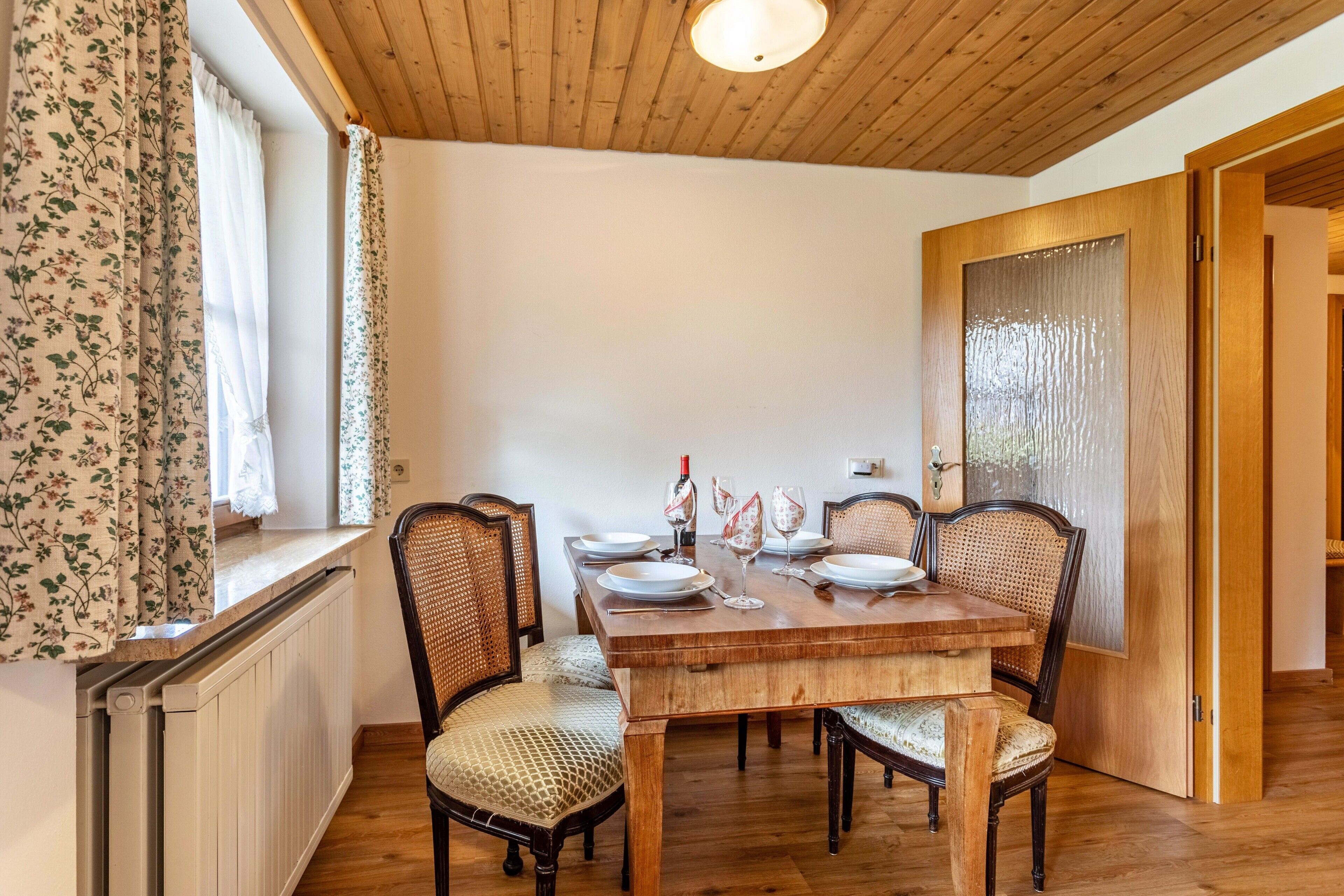 Een eetkamer met een houten tafel voor vier personen, omringd door rieten stoelen, een radiator en een raam met bloemen gordijnen.