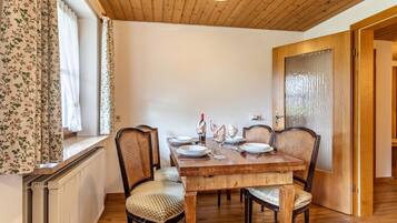 Een eetkamer met een houten tafel voor vier personen, omringd door rieten stoelen, een radiator en een raam met bloemen gordijnen.