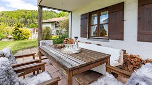 A wooden patio with a table and chairs, a window with shutters, and a view of a green landscape. - Holiday Home Bergblick for 4 Guests (Unterwössen)