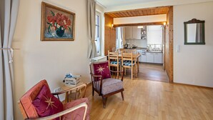 Una habitación con piso de madera, una mesa de comedor con sillas, un radiador, una pequeña mesa con una caja y un cuadro en la pared.