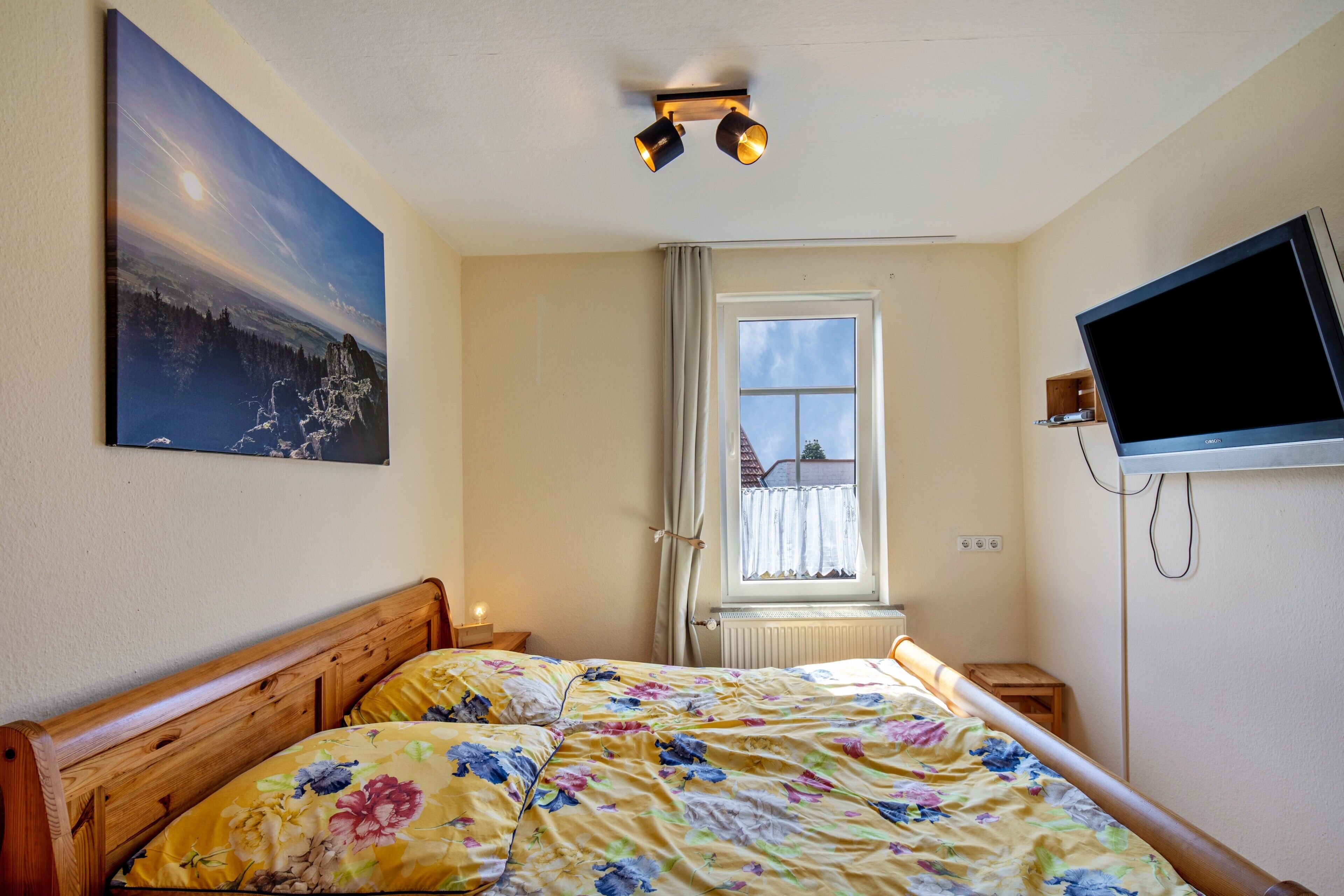 A bedroom with a bed, a TV, a window, and a painting on the wall.