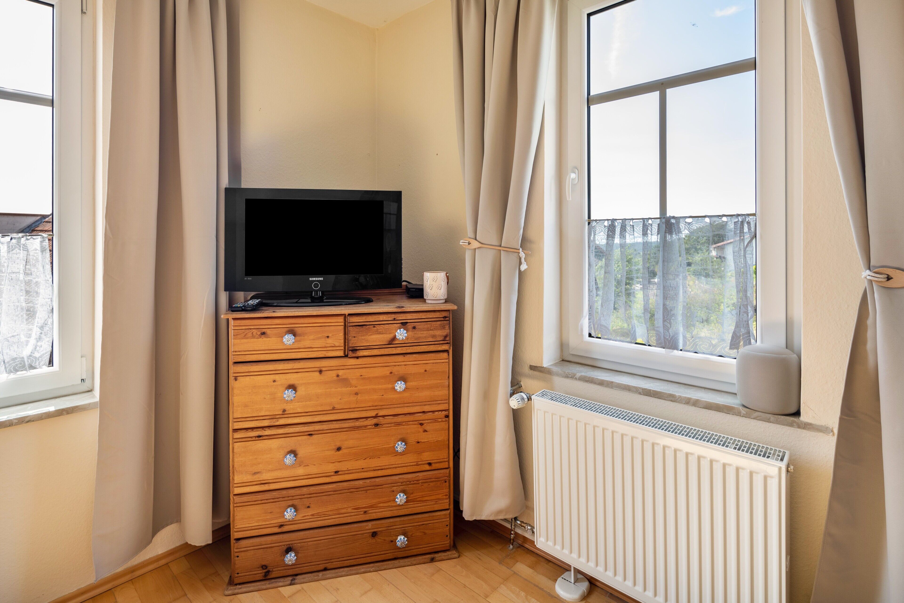 Una habitación con un aparador de madera, un televisor encima, una ventana con cortinas y un radiador.