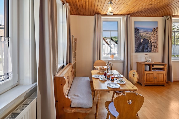 A room with a wooden bench, a dining table with chairs, a radiator, a window with curtains, and a painting on the wall.