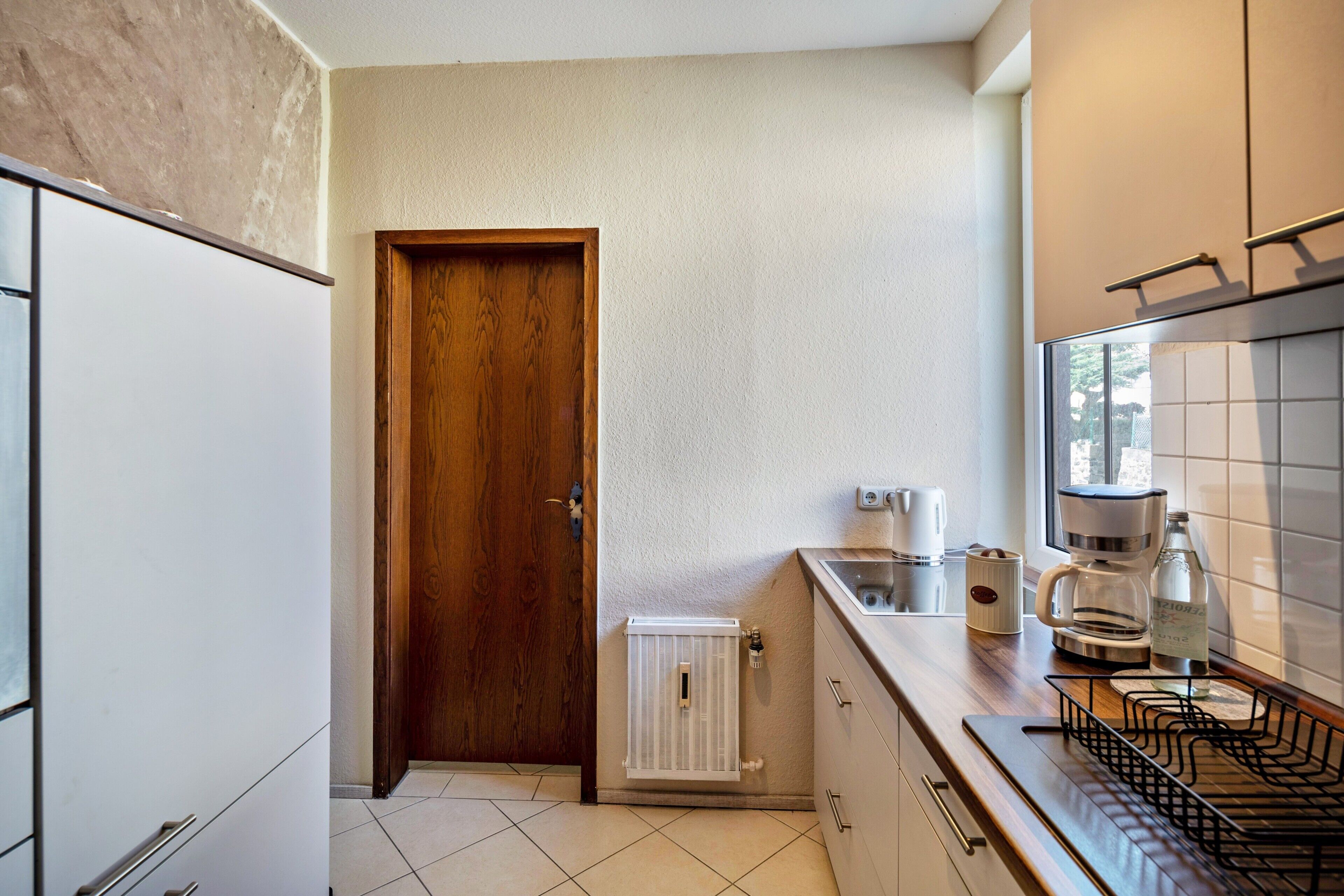 A compact kitchen with a wooden door, white cabinets, a coffee maker, and a kettle on the counter.