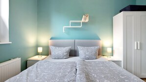 A bedroom with a bed, two bedside tables, a nightstand, a wardrobe, and a wall-mounted shelf.