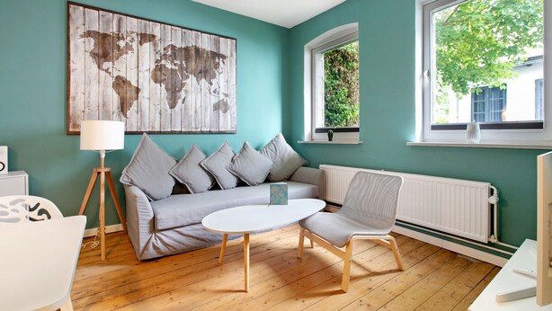 A room with a teal wall, a world map artwork, a white sofa, a small round table, a white chair, and a window with a view of greenery.