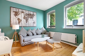 A room with a teal wall, a world map artwork, a white sofa, a small round table, a white chair, and a window with a view of greenery.