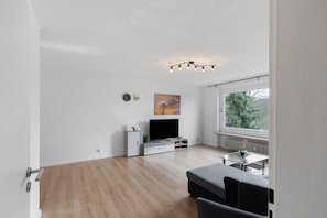 Una sala moderna con televisor de pantalla plana, una mesa de centro de vidrio, un sofá y una ventana con vistas a árboles.