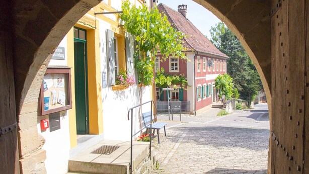 A cobblestone street with a building entrance framed by an archway, a bench, and a small tree with hanging flowers. - Apartment 'Ferienwohnung Kammer' with Private Terrace, Shared Garden and Wi-Fi (Iphofen)