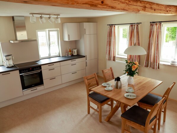 A modern kitchen with white cabinets, a built-in oven, and a coffee maker. There is a dining table with chairs and a vase of flowers.