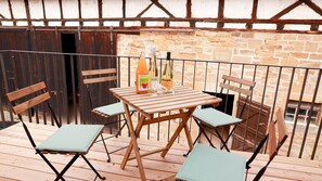 Une table en bois, entourée de chaises, sur un balcon dont les murs sont en pierre et qui est agrémenté d’une rambarde en métal.