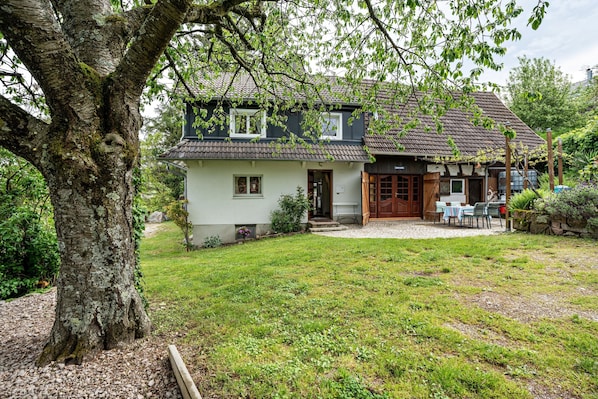 Une maison à deux étages avec un porche à l’avant, un petit jardin et un grand arbre au premier plan.