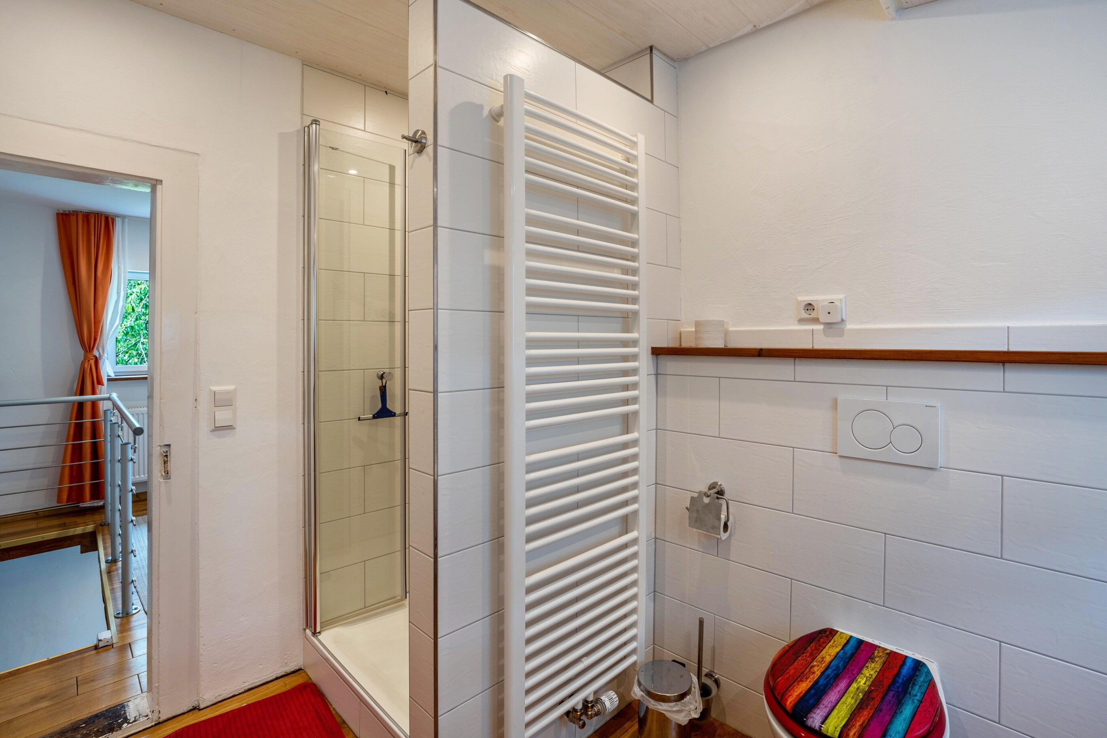 Un baño con cabina de ducha de vidrio, un perchero blanco, un inodoro con asiento de colores y suelo de madera.