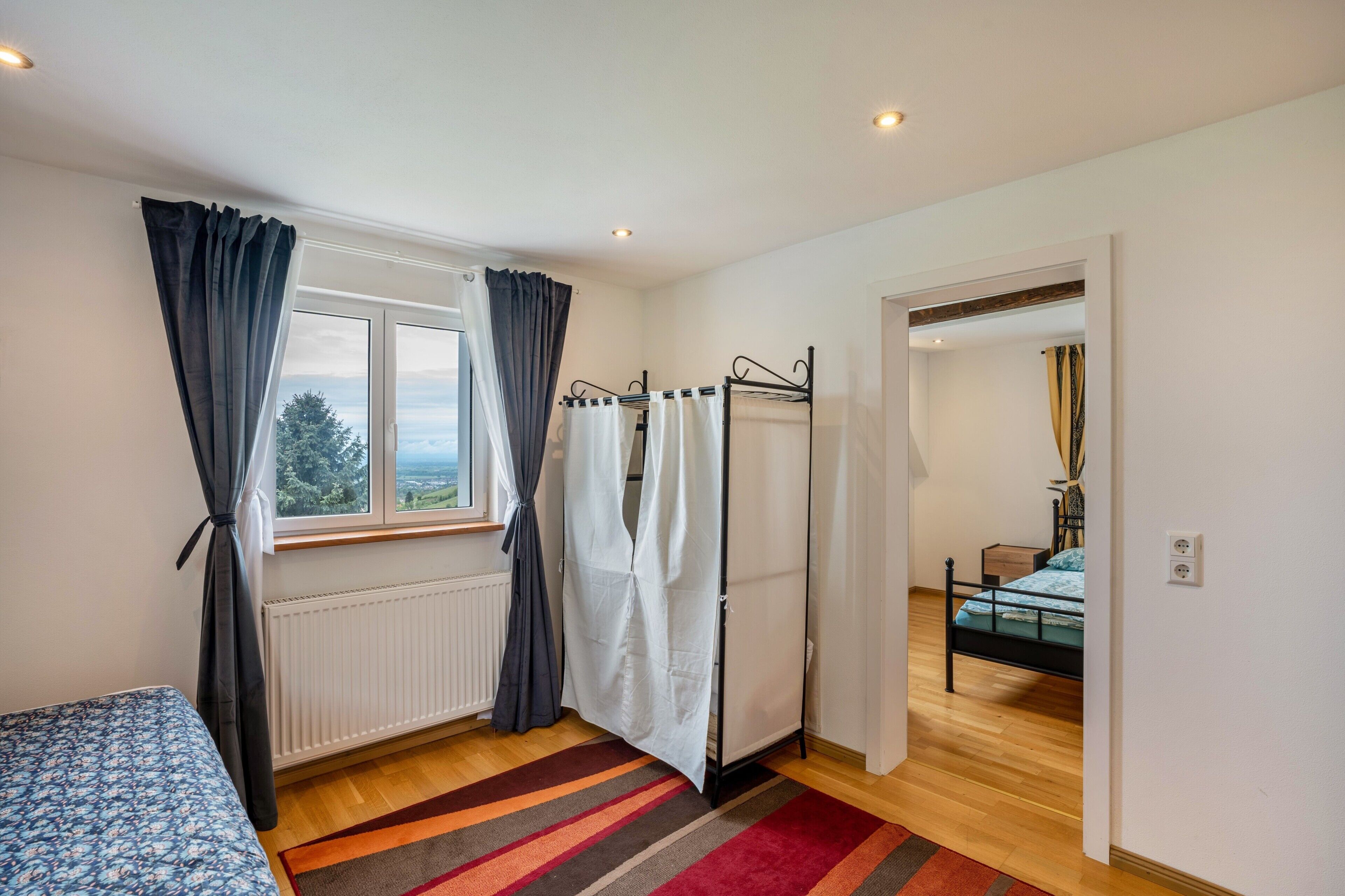 A bedroom with a bed, a window with curtains, a radiator, a striped rug, and a door leading to another room.