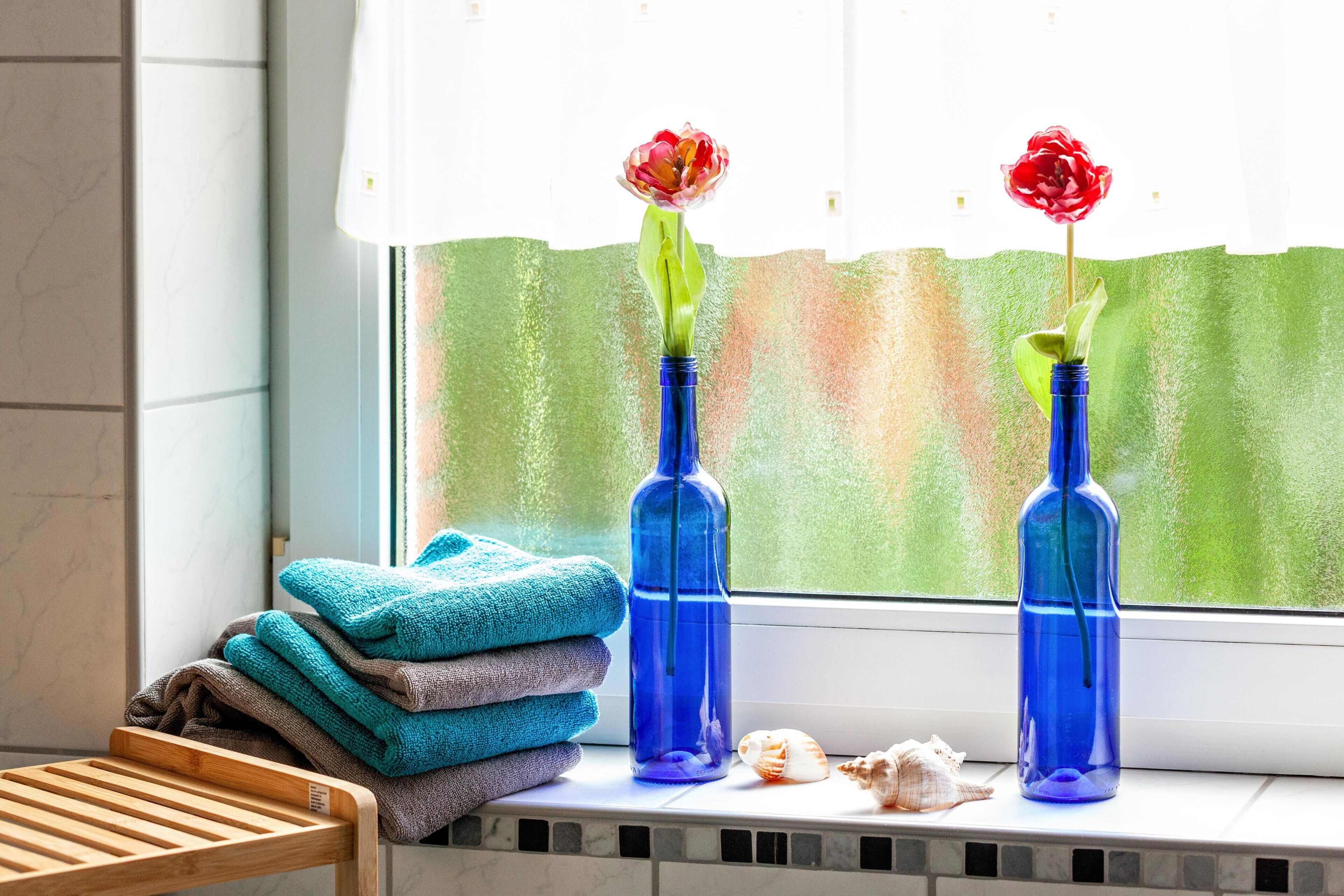 Una ventana con dos jarrones azules con flores rojas, una pila de toallas y conchas marinas en el alféizar.