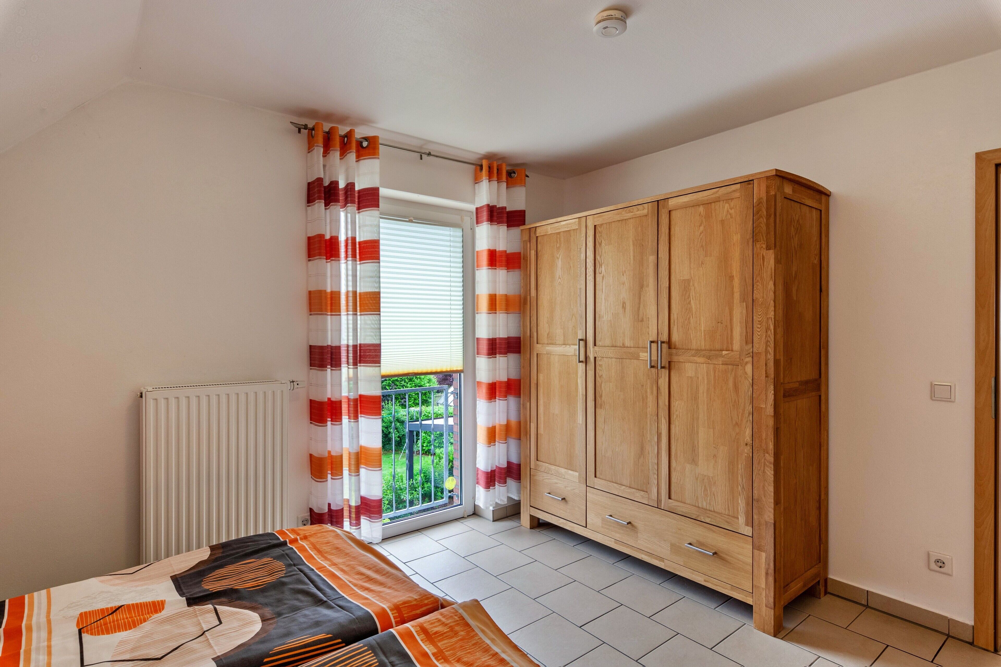 Ein Schlafzimmer mit Bett, Schrank, Heizkörper und einem Fenster mit Vorhängen.