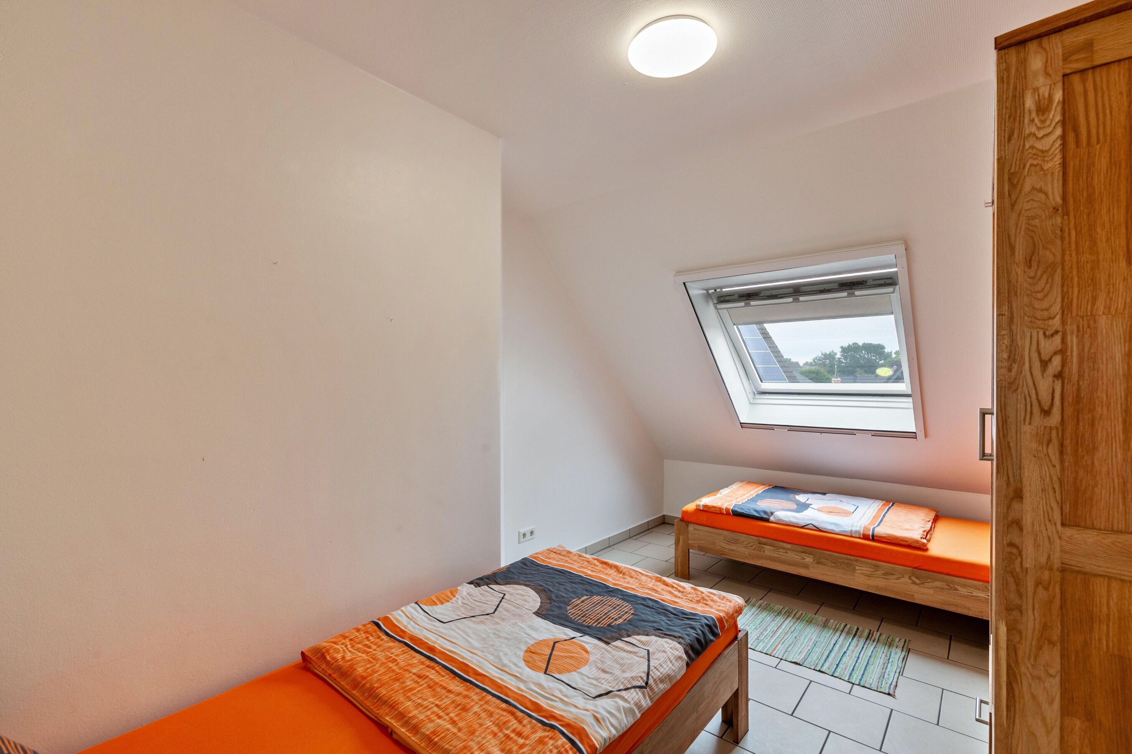 Ein kleines Zimmer mit einem Einzelbett, schrägen Wänden, einem Fenster und einem Holzschrank.