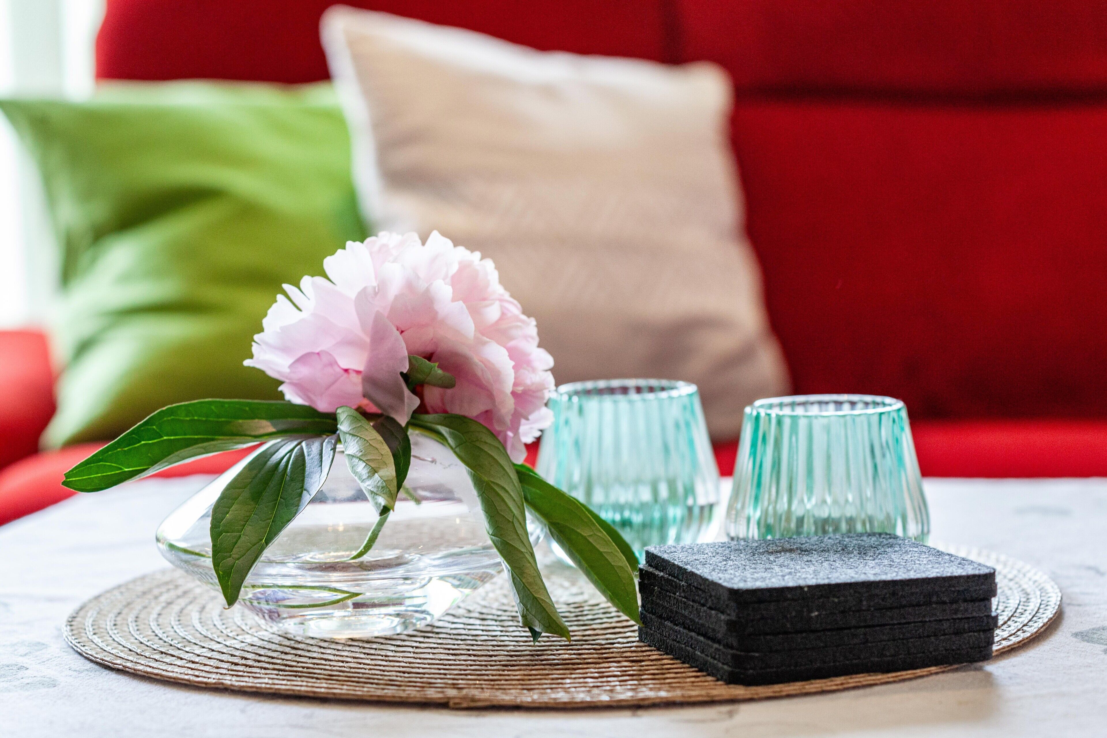 A clear glass vase with a pink flower, two glass tumblers, and a stack of black coasters on a woven placemat.