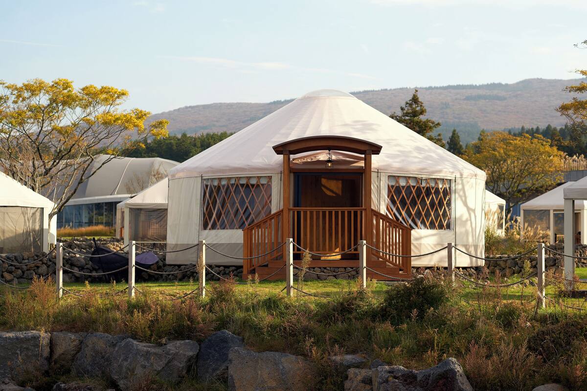 A yurt with a wooden porch and lattice windows, surrounded by a chain-link fence and set in a grassy area with mountains in the background.