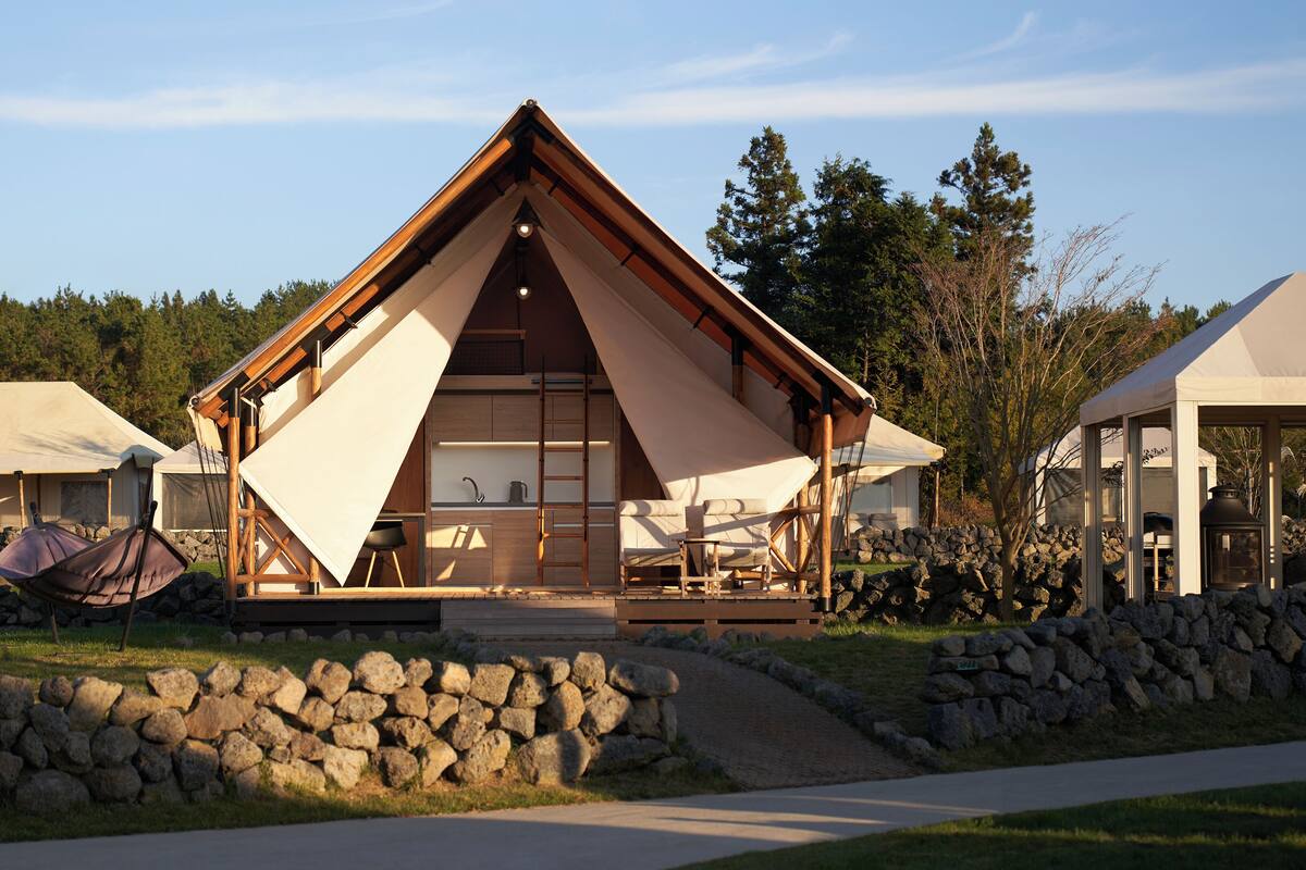 A modern, triangular-roofed cabin with a stone wall entrance, a small porch, and a view of the surrounding forest.