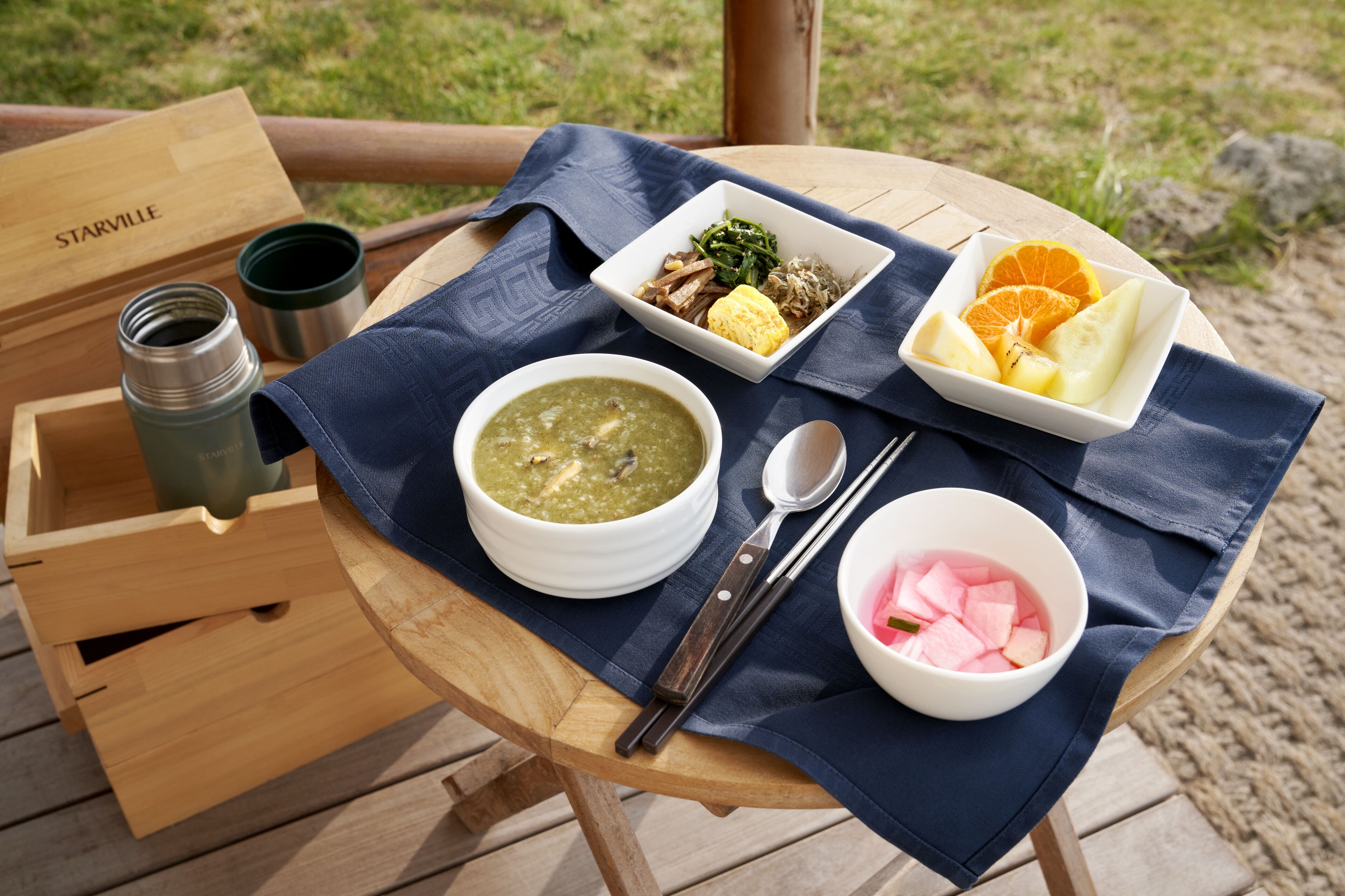 A wooden table with a blue cloth, a bowl of soup, a plate with vegetables and meat, a bowl of fruit, and a wooden box labeled "Starville".