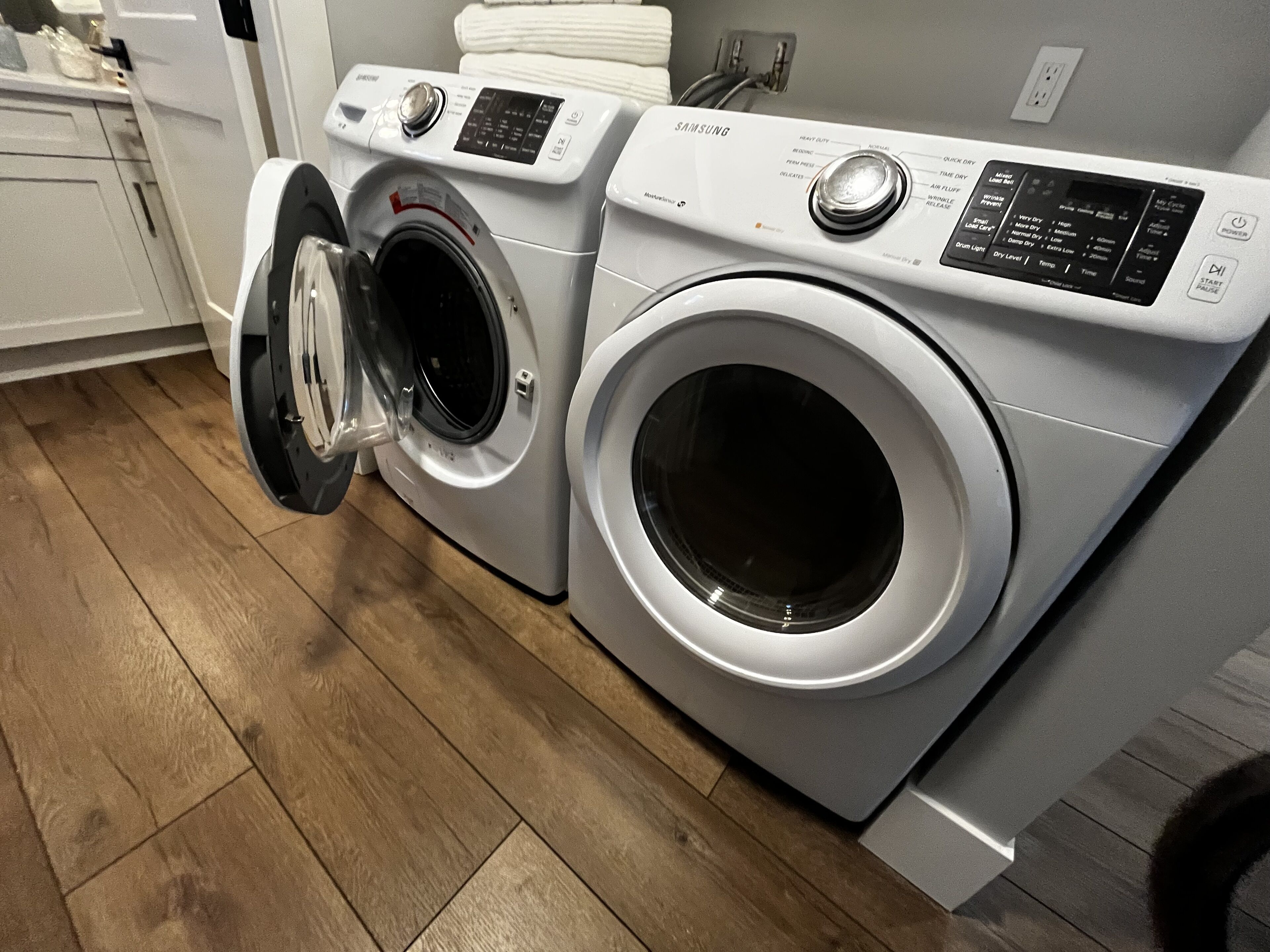 Un lave-linge et un sèche-linge à chargement frontal, installés dans une buanderie dont le sol est recouvert de parquet.