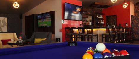 A pool table with billiard balls, a television mounted on the wall, and a bar area with chairs and shelves.