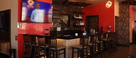 A bar area with a long counter, multiple bar stools, a television screen, and a kitchen in the background.