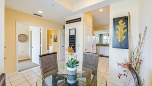 A dining area with a glass table surrounded by wicker chairs, a clock on the wall, and a decorative plant. - Naples Resort Condo on 2 Championship Golf Courses (Naples)