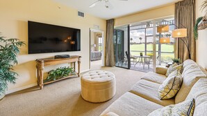 A living room with a large flat-screen TV, a round ottoman, a sofa with patterned cushions, and a ceiling fan. - Naples Resort Condo on 2 Championship Golf Courses (Naples)