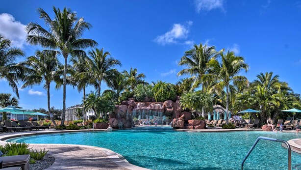 A swimming pool with palm trees and lounge chairs, surrounded by a paved area and a waterfall feature. - Naples Resort Condo on 2 Championship Golf Courses (Naples)