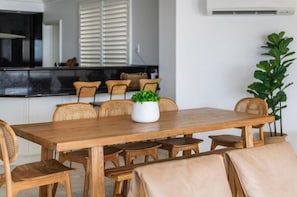 A dining area with a wooden table and chairs, a potted plant, and a kitchen in the background. - Oceans 8 - Oceanstays -Absolute Oceanfront with Fireplace (Yamba)